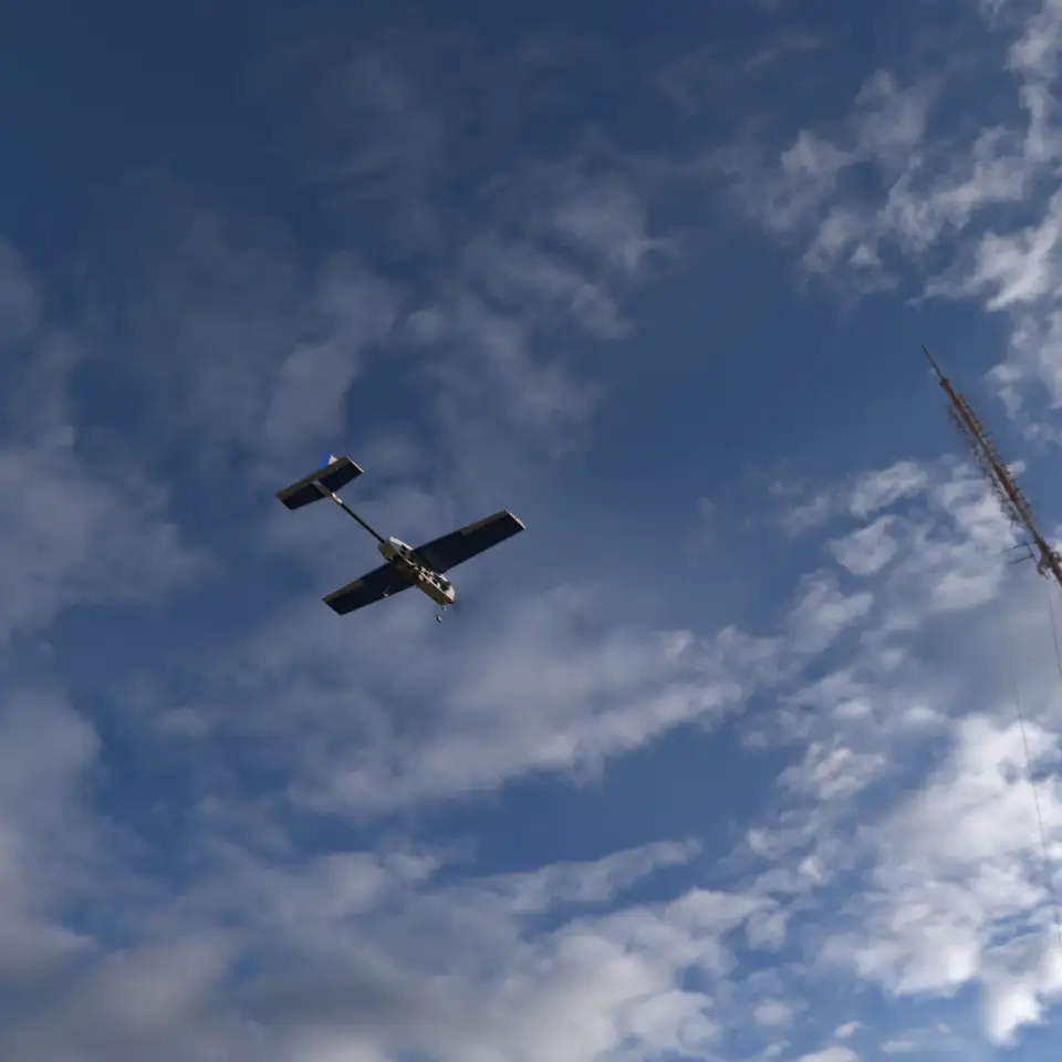Photorealistic render of the Teknofest fixed-wing UAV in flight, seen from below against a cloudy blue sky beside a radio mast.