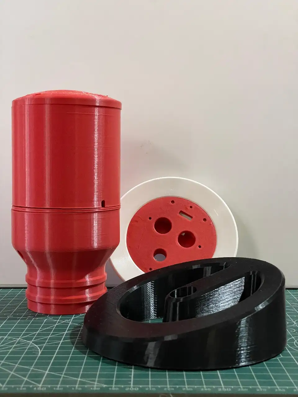 Three 3D-printed prototype parts in red, white and black on a cutting mat.